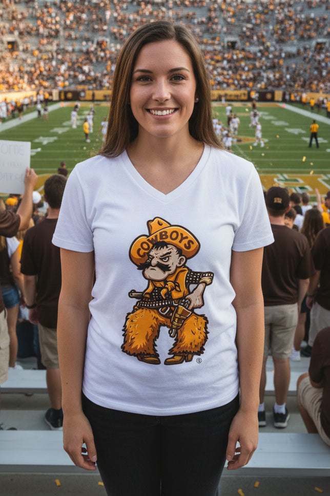 A smiling woman stands in a stadium wearing a A white women's V-neck t-shirt featuring University of Wyoming Cowboy Joe graphic on a white fabric background. The cowboy wears a yellow hat with the word 'COWBOYS' and a matching bandana, with yellow furry chaps and boots, holding a gun. The background shows a football field and crowd in the stands.