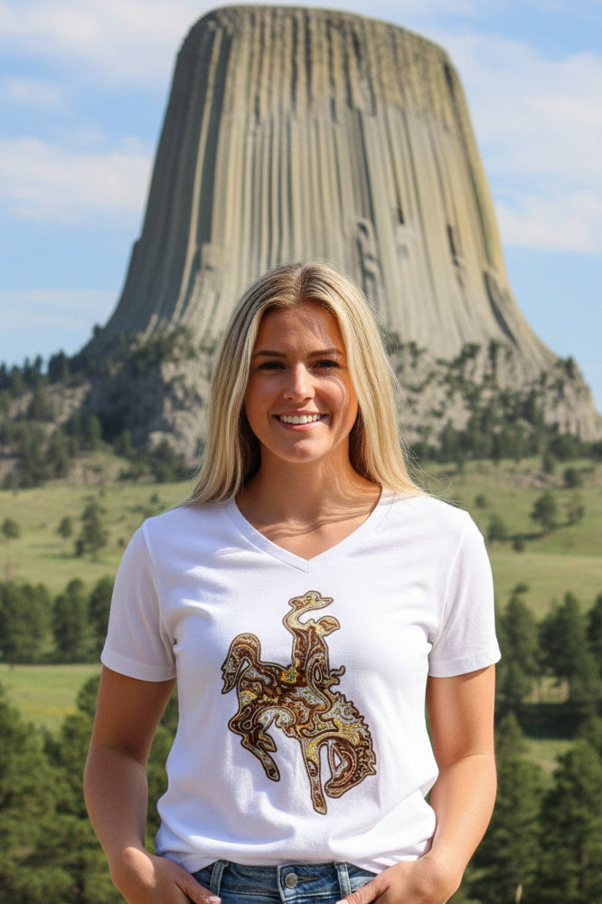 A smiling blonde woman stands outdoors in front of a Devils Tower, wearing a white V-neck T-shirt with a brown and gold bucking horse graphic on the front.
