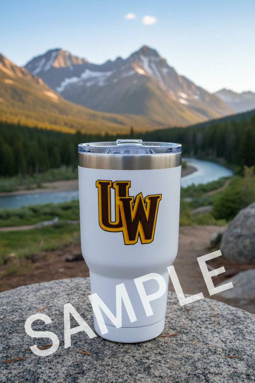 A white insulated travel tumbler with a clear lid and a large yellow and brown 'UW' logo on the front, set on a stone surface with a mountain and river landscape in the background.