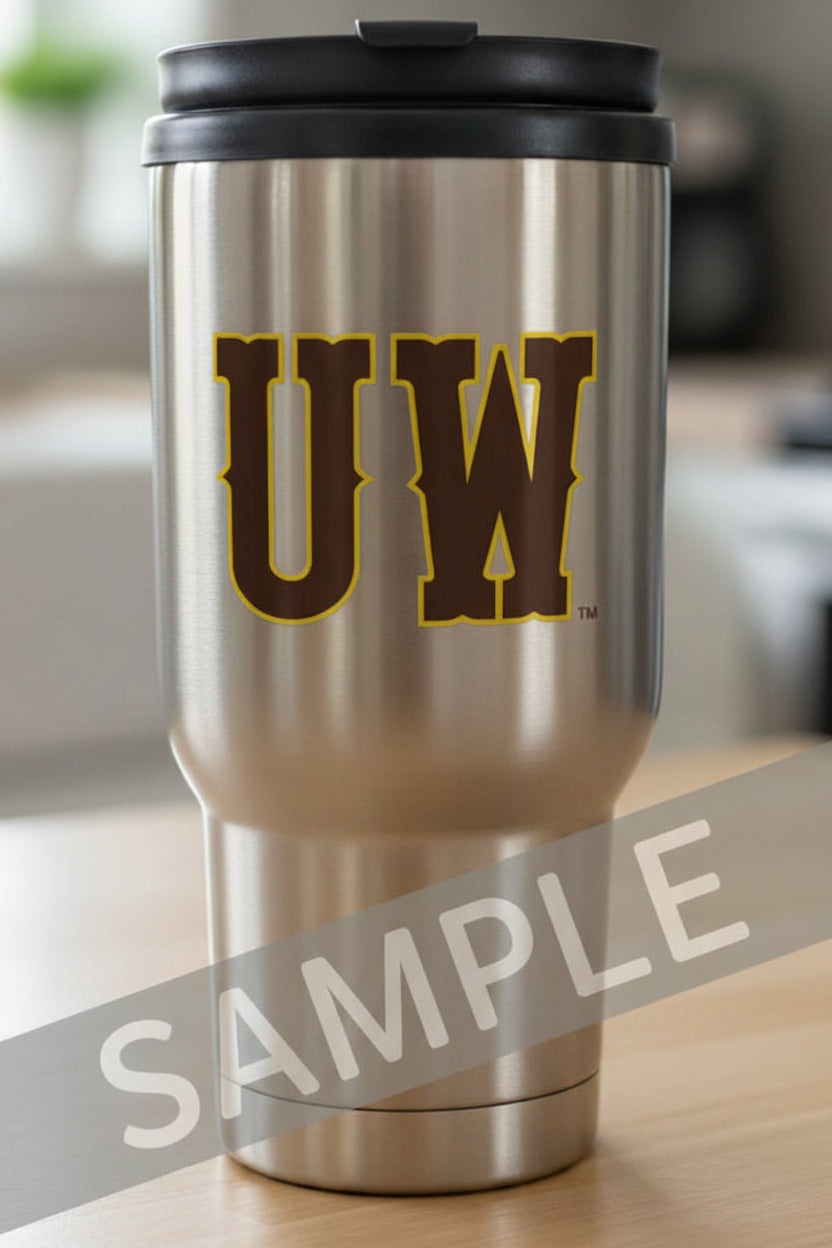 A stainless steel insulated travel tumbler with a black lid, featuring a large brown and gold 'UW' logo on the front, sitting on a light wood surface with a blurred indoor background.