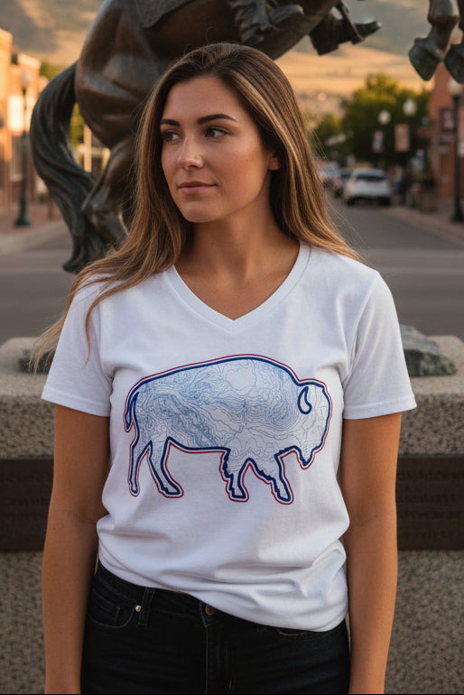 A woman wearing a white V-neck t-shirt with a bison outline filled with a map pattern and outlined in red and blue, standing outdoors in front of a bronze horse statue.