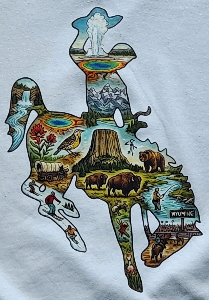 A close-up of a white T-shirt featuring a colorful graphic print of the Wyoming Bucking Horse logo, filled with illustrated scenes of wildlife, mountains, a geothermal pool, a covered wagon, and outdoor activities.