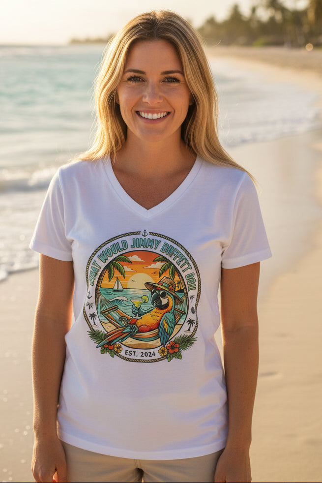 A smiling blonde woman stands on a sandy beach wearing a white V-neck t-shirt with a colorful tropical graphic featuring a parrot, sunset, and the text 'What would Jimmy Buffet do!'.