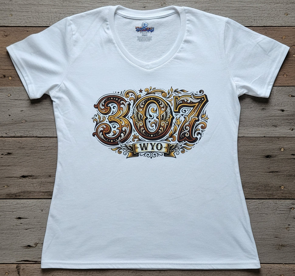 A white short-sleeve V-neck T-shirt laid flat on a rustic wooden plank background, featuring a large decorative graphic on the front with the numbers '307' in ornate brown, gold, and white lettering above a banner that reads 'WYO'.