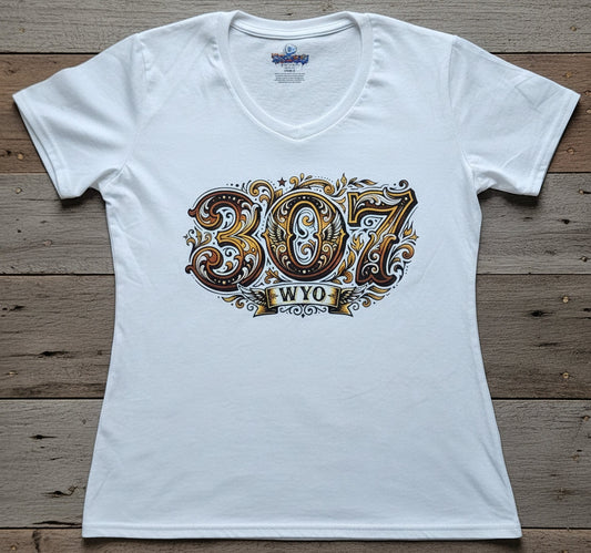 A white short-sleeve V-neck T-shirt laid flat on a rustic wooden plank background, featuring a large decorative graphic on the front with the numbers '307' in ornate brown, gold, and white lettering above a banner that reads 'WYO'.