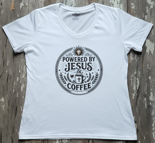 A white women's V-neck T-shirt laid flat on a weathered wood surface, featuring a circular brown graphic print on the chest that reads 'POWERED BY JESUS & COFFEE' with a cross and coffee cup motif.