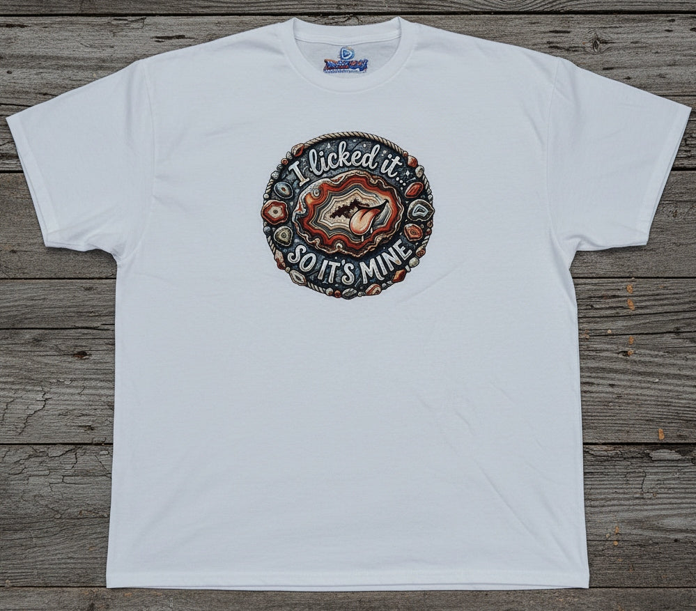 A white short-sleeve t-shirt laid flat on a rustic gray-brown wooden plank background. The shirt features a circular graphic print on the chest with a cartoon agate and the text 'I licked it SO IT'S MINE'.