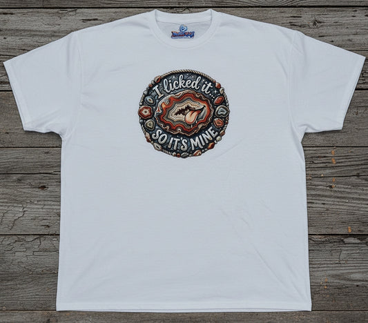 A white short-sleeve t-shirt laid flat on a rustic gray-brown wooden plank background. The shirt features a circular graphic print on the chest with a cartoon agate and the text 'I licked it SO IT'S MINE'.