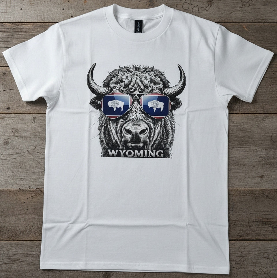 A white short-sleeve crewneck t-shirt laid flat on a rustic wooden background. The shirt features a graphic print of a bison wearing sunglasses with white bison silhouettes inside, and the word 'WYOMING' printed below.