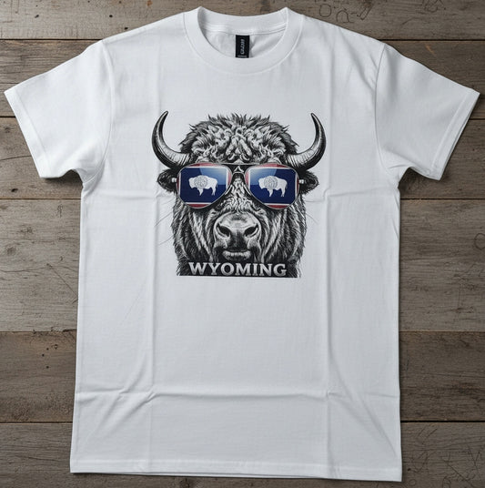 A white short-sleeve crewneck t-shirt laid flat on a rustic wooden background. The shirt features a graphic print of a bison wearing sunglasses with white bison silhouettes inside, and the word 'WYOMING' printed below.