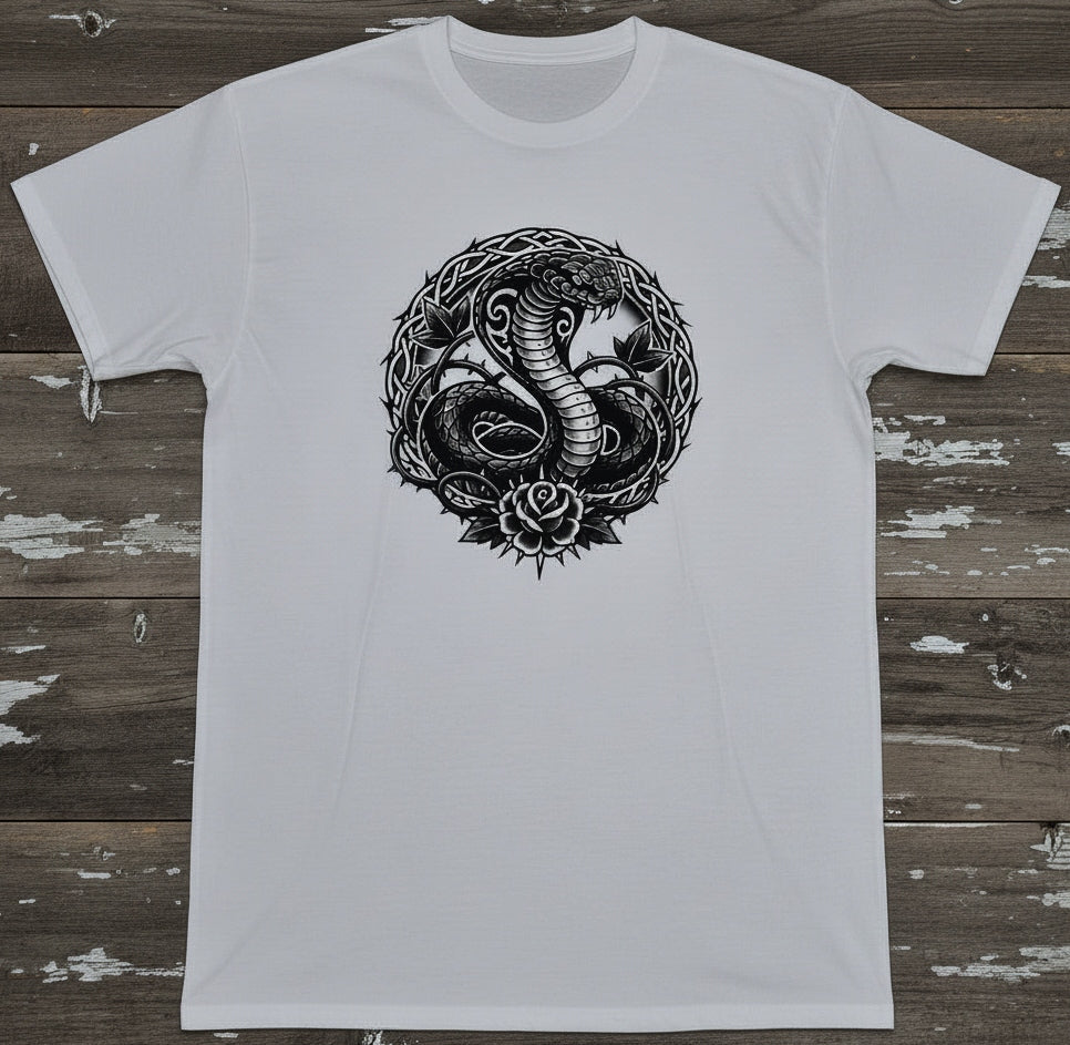 A white short-sleeve crewneck t-shirt featuring a detailed black graphic of a coiled cobra snake inside a circular frame with a rose at the bottom, laid flat on a rustic, weathered wooden plank background.