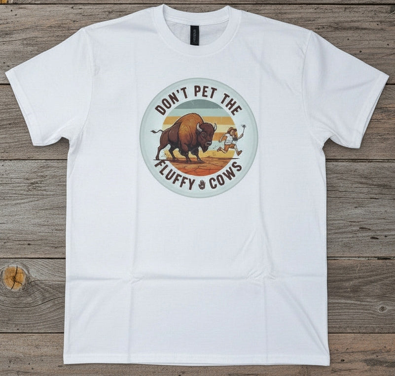 A white short-sleeve crewneck t-shirt laid flat on a rustic wooden plank background. The shirt features a circular graphic in the center with a bison, tourist, and the text 'DON'T PET THE FLUFFY COWS'.