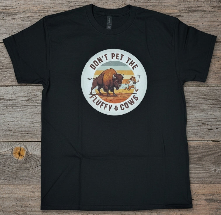 A black short-sleeve t-shirt laid flat on a rustic wooden surface, featuring a circular graphic on the chest with a bison, tourist, and the text 'DON'T PET THE FLUFFY COWS'.