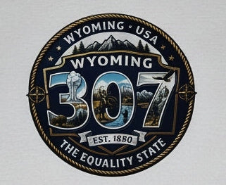 A white short-sleeve crewneck t-shirt laid flat on a dark wood plank background, featuring a circular graphic on the chest with the text 'WYOMING 307 THE EQUALITY STATE' and a mountain illustration.