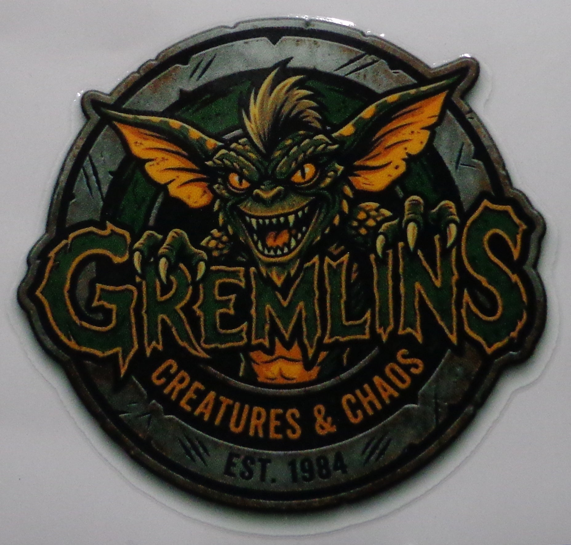 A circular Gremlins-themed sticker  featuring a green gremlin, above the word 'GREMLINS' in stylized lettering. The design includes the phrases 'CREATURES & CHAOS' and 'EST.