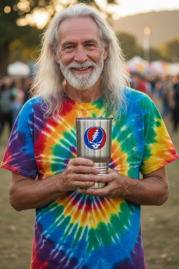 A smiling man with long white hair and a beard holds a stainless steel tumbler with a Grateful Dead Steal the Night skull logo. He is wearing a vibrant rainbow tie-dye t-shirt in an outdoor festival setting.