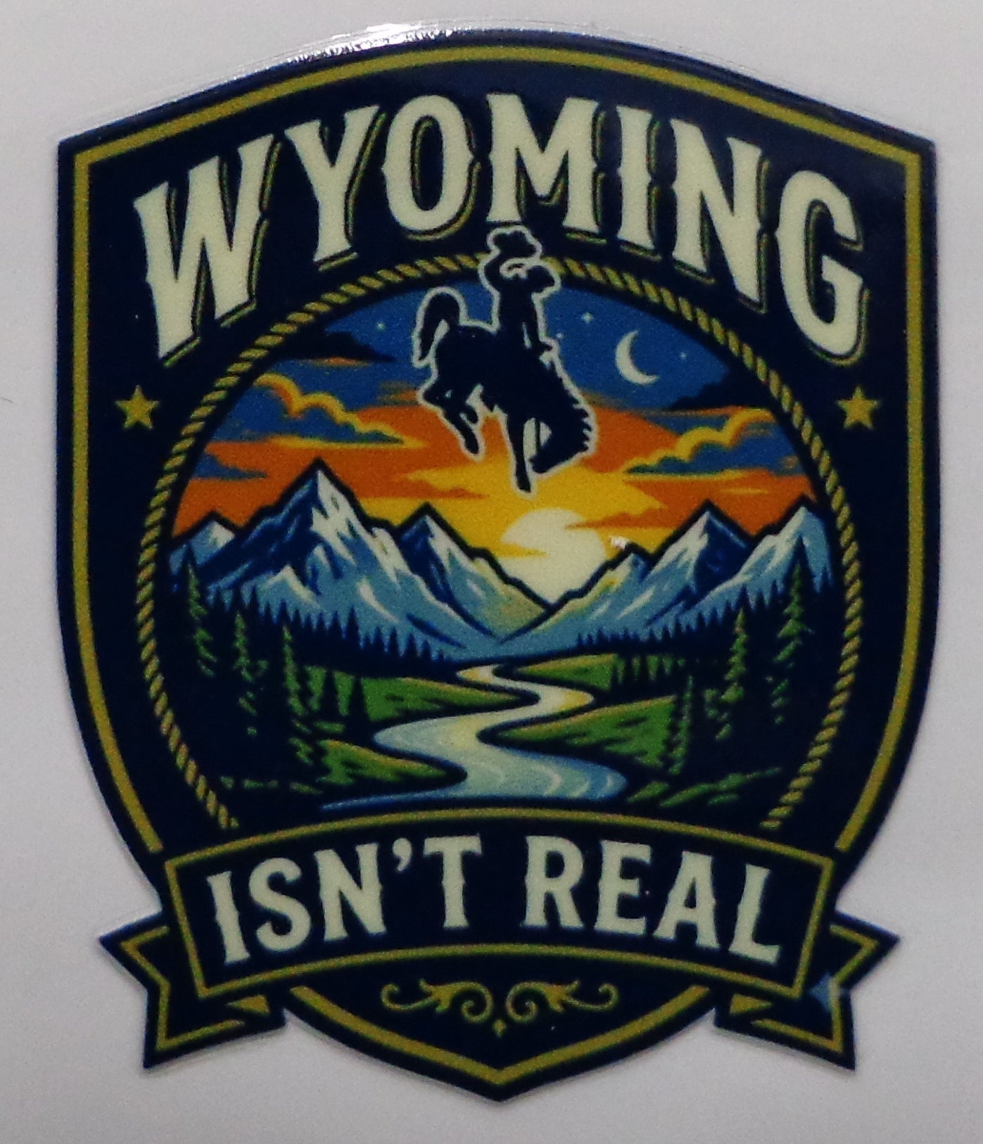 A shield-shaped sticker featuring the words “WYOMING ISN’T REAL” around a western mountain landscape scene with a cowboy riding a bucking horse at sunset.