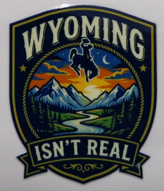 A shield-shaped sticker featuring the words “WYOMING ISN’T REAL” around a western mountain landscape scene with a cowboy riding a bucking horse at sunset.