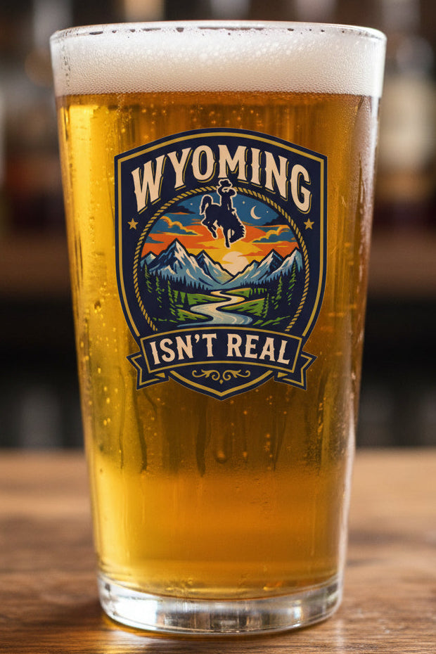 A shield-shaped sticker featuring the words “WYOMING ISN’T REAL” around a western mountain landscape scene with a cowboy riding a bucking horse at sunset, placed on a beer glass