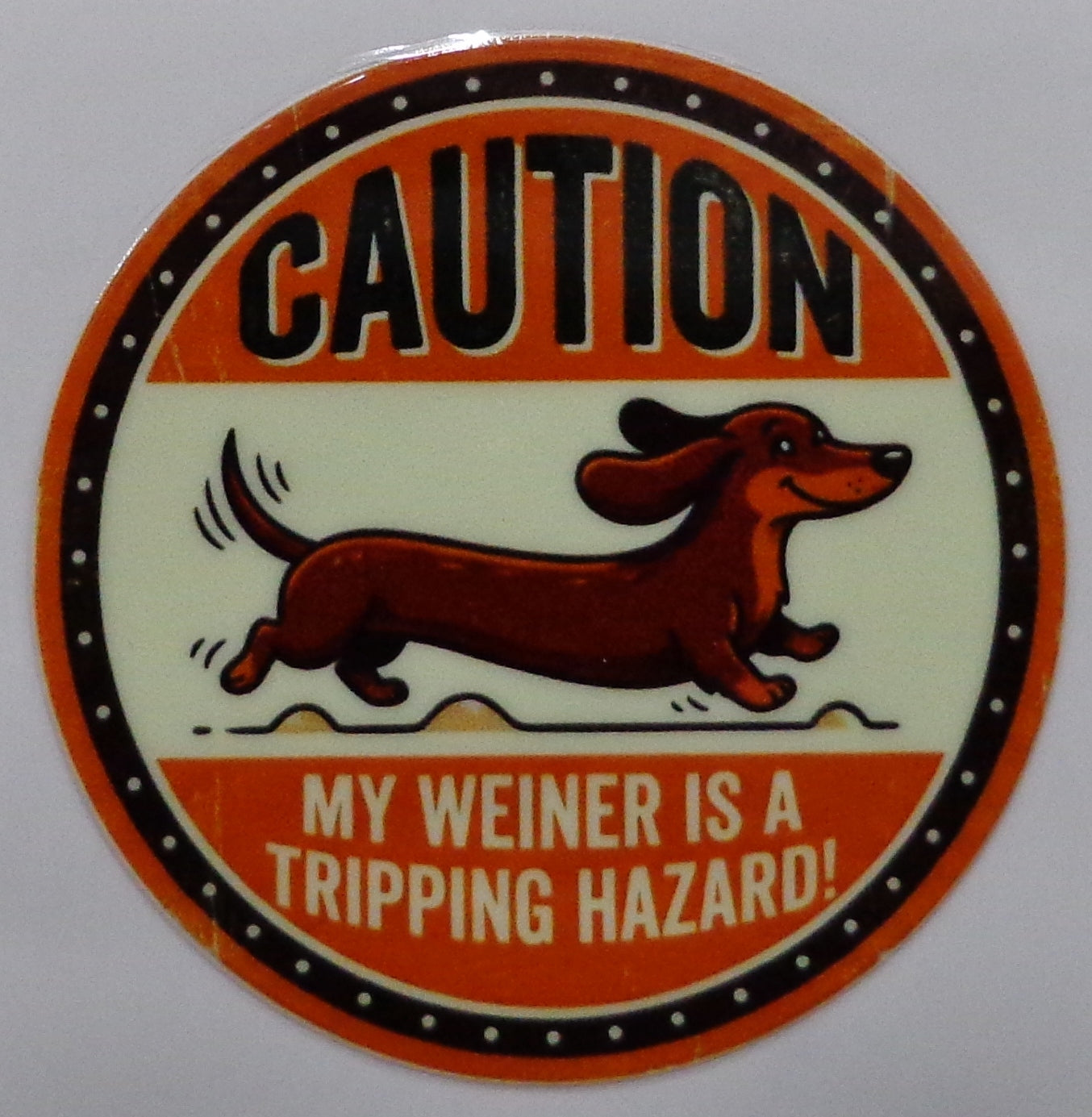 A circular novelty sticker featuring a cartoon dachshund and the text 'CAUTION MY WEINER IS A TRIPPING HAZARD!' in orange, black, and cream colors.