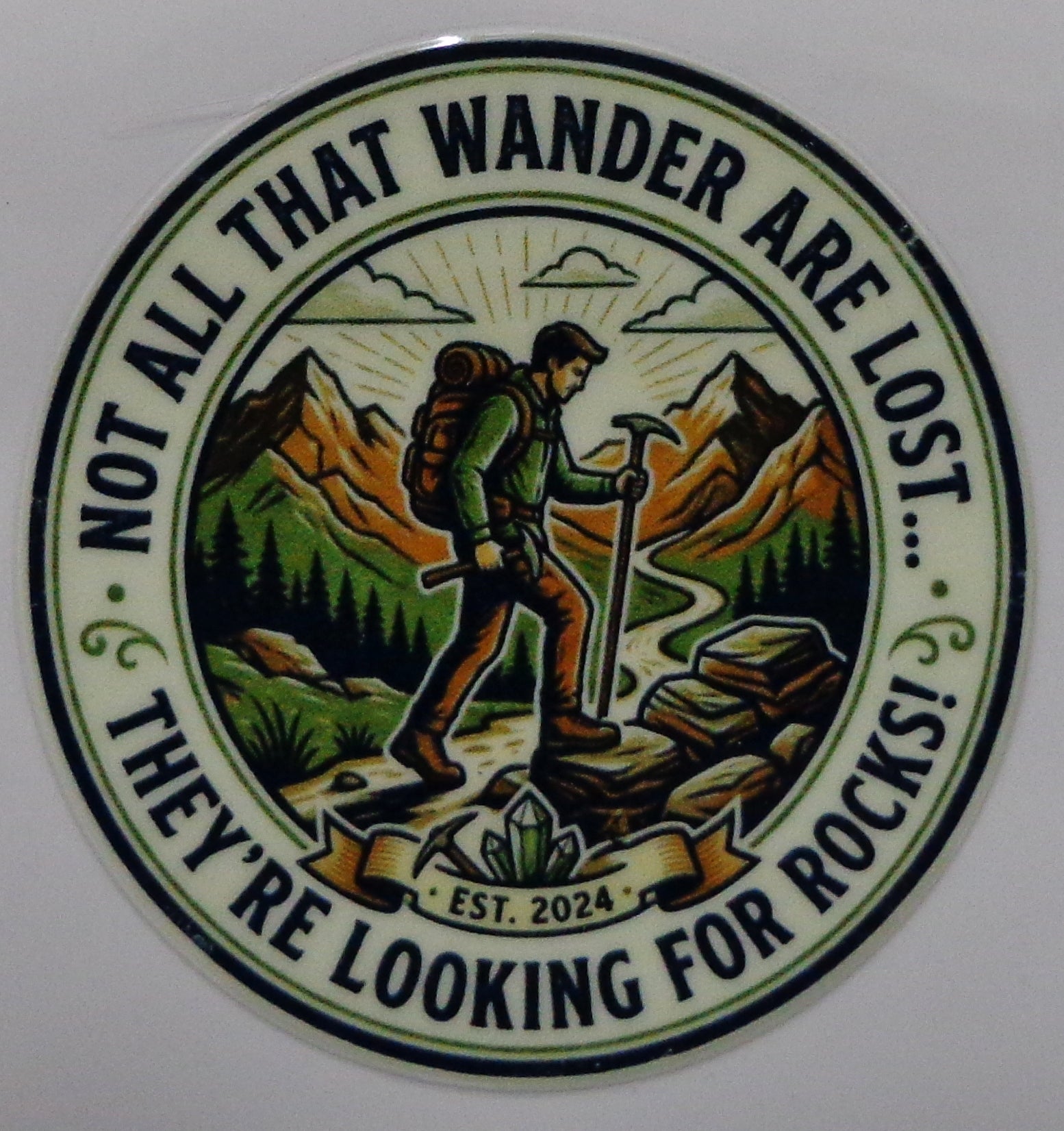 A circular vintage-style sticker or decal featuring a hiking man with a backpack and trekking pole in front of mountains, with the text 'NOT ALL THAT WANDER ARE LOST... THEY'RE LOOKING FOR ROCKS.