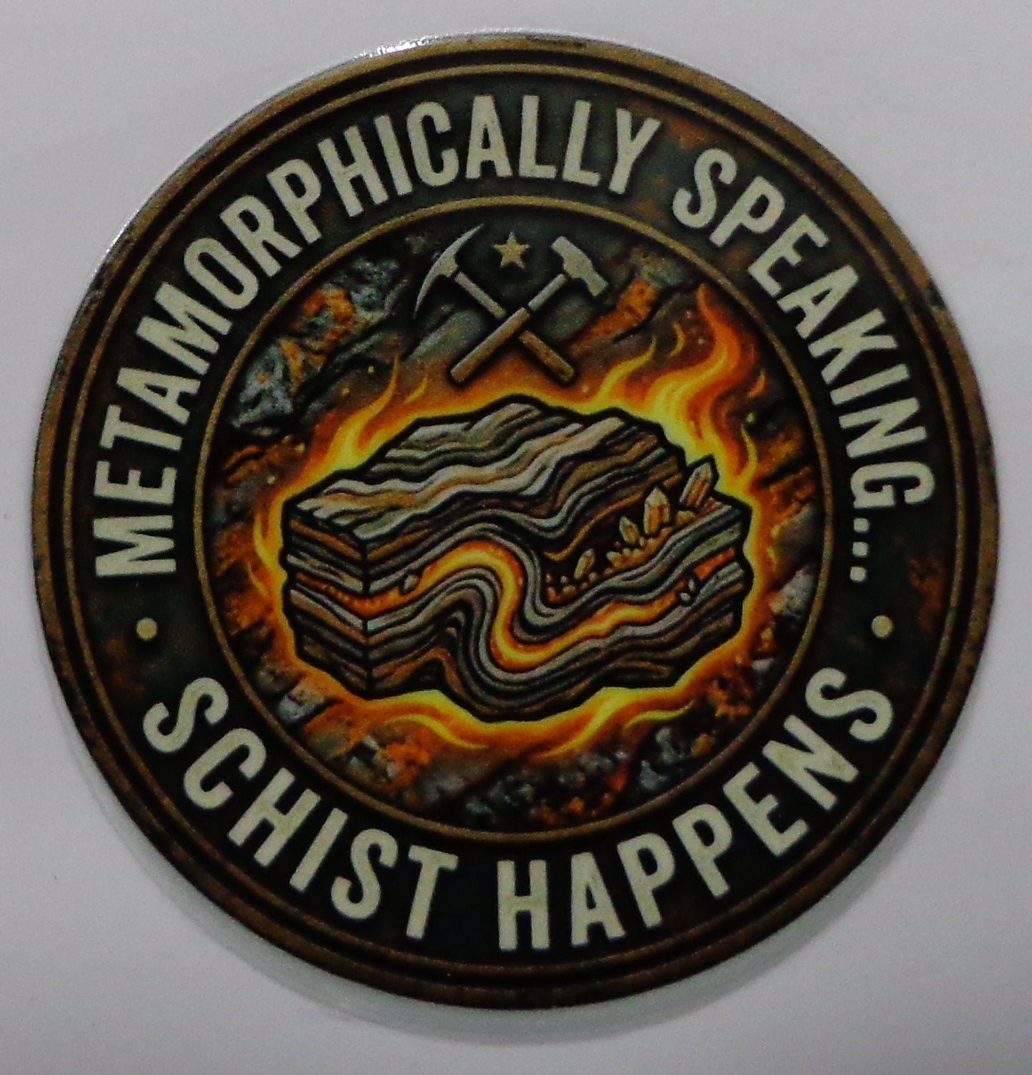 A circular sticker featuring a stylized illustration of layered rock formations with crossed picks and a star, surrounded by the text 'METAMORPHICALLY SPEAKING.... SCHIST HAPPENS'.