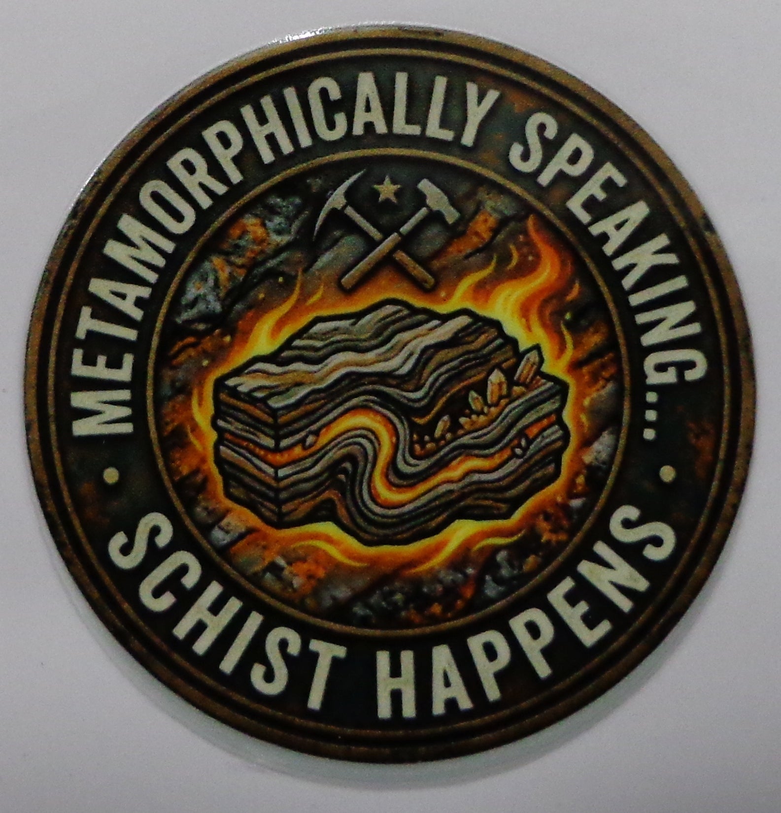 A circular sticker featuring a stylized illustration of layered rock formations with crossed picks and a star, surrounded by the text 'METAMORPHICALLY SPEAKING.... SCHIST HAPPENS'.