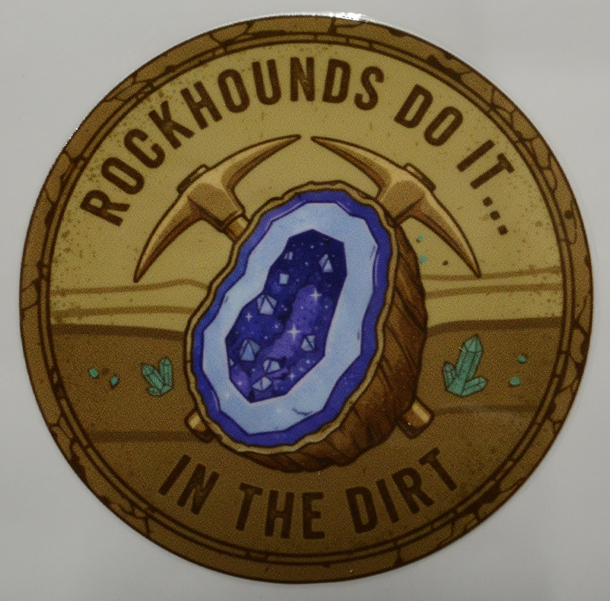 Circular sticker with a geode and pickaxe graphic, text 'Rockhounds do it... in the dirt', on a beige background.